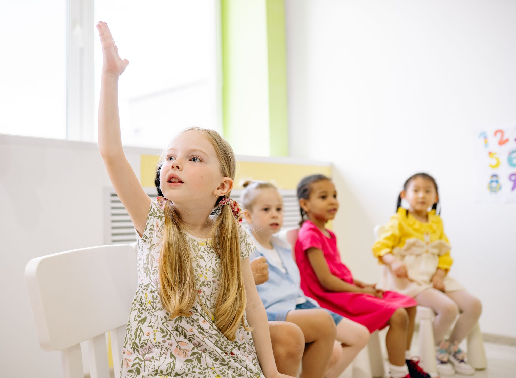 Children in a bright, sunlit classroom; a girl raises her hand beside a large window.